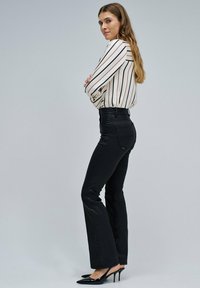 Mujer de pie con los brazos cruzados, vistiendo una blusa blanca a rayas, pantalones negros de cintura alta y tacones negros de punta contra un fondo gris.