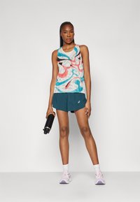 Sport tanktopp med flerfärgad abstrakt mönster, tillsammans med turkosa shorts. Svart vattenflaska hålls i vänster hand, vita strumpor och rosa sneakers.