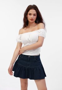 Weißes Off-Shoulder-Oberteil aus strukturiertem Stoff mit Puffärmeln, kombiniert mit einem dunklen, taillierten Jeansrock mit ausgestelltem Schnitt, der einen volutenförmigen Saum und Taschen hat.