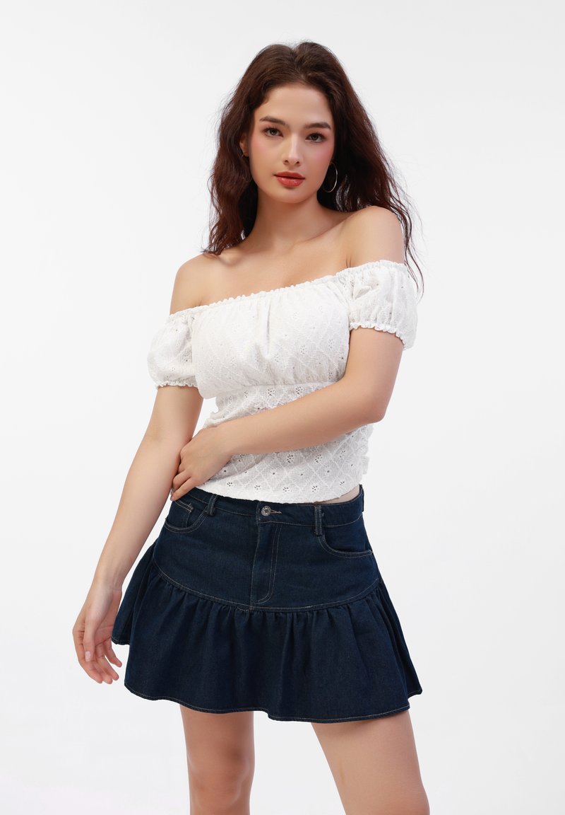 Weißes Off-Shoulder-Oberteil aus strukturiertem Stoff mit Puffärmeln, kombiniert mit einem dunklen, taillierten Jeansrock mit ausgestelltem Schnitt, der einen volutenförmigen Saum und Taschen hat.