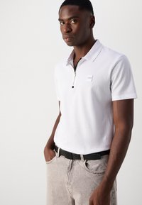 Hvid polo T-shirt med en lynlåskrave, der har et lille logo patch. Parret med lysegrå denimbukser og et sort bælte.