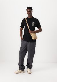 Schwarzes T-Shirt mit Logo, graue Cargo-Hosen mit Seitentaschen und beiger Umhängetasche. Die Schuhe sind hellbeige und für Komfort und Unterstützung ausgelegt.
