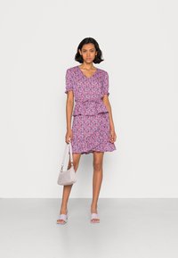 Vestido com padrão floral em rosa e roxo, com mangas curtas, cintura peplum e barra com babados. Os acessórios incluem uma mala lilás e sapatilhas.