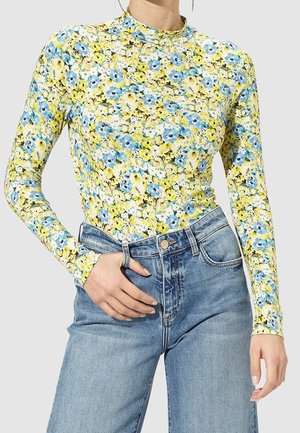 Kobieta w dopasowanym, długim rękawie bluzce w kwiatowy wzór w kolorach niebieskim i żółtym, ubrana w wysokowodne jasnoniebieskie dżinsy, na jednolitym tle.