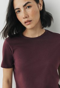 Bordeaux kortærmet t-shirt lavet af blødt stof, med rund halsudskæring og en glat overflade, båret af en model med mørkt hår.