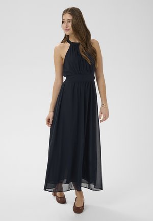 Vrouw met lang haar, gekleed in een mouwloze marineblauwe maxi-jurk met halternek en bruine gevlochten platte schoenen, staand tegen een effen achtergrond.