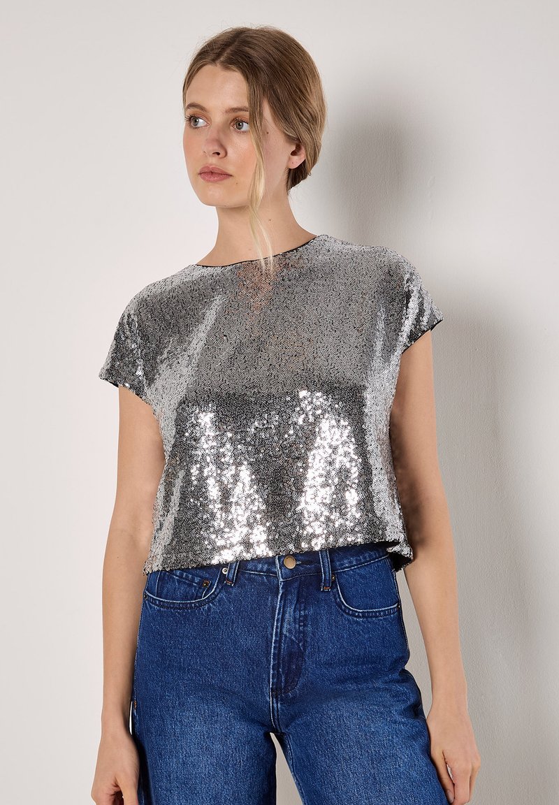 Silberner, mit Pailletten besetzter Kurzarm-Top mit Rundhalsausschnitt, kombiniert mit blauen, hoch taillierten Jeans in einem lässigen, cropped Design.