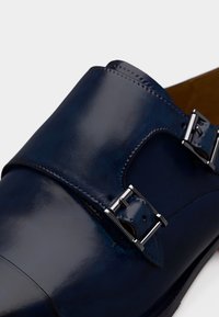 Σκούρο μπλε δερμάτινο παπούτσι διπλού μονκ με ασημένιες αγκράφες και λείο γυαλιστερό φινίρισμα σε λευκό φόντο.