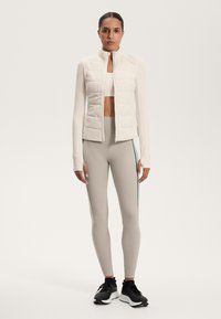 Light-weight padded vest in cream, form-fitting long-sleeve top, and beige leggings with teal side stripes, paired with black athletic shoes.