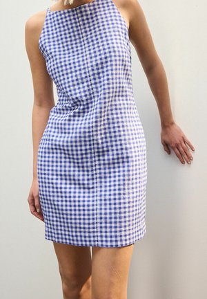 Femme portant une robe ajustée sans manches à carreaux bleus et blancs, debout devant un fond blanc uni.
