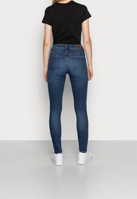 Jeans oscuros ajustados de talle medio, con bolsillos traseros que presentan un sutil bordado, combinados con zapatillas blancas sobre un fondo liso.