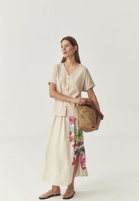 Haut beige clair à manches courtes et boutons, associé �à une jupe maxi crème ornée de motifs floraux colorés et d'une fente latérale. Tient un sac en osier.