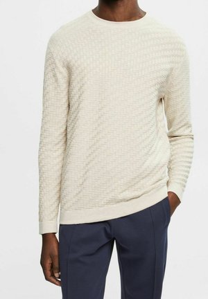 Uomo che indossa un maglione a maniche lunghe color crema con trama e pantaloni blu navy, mani rilassate lungo i fianchi su uno sfondo semplice.