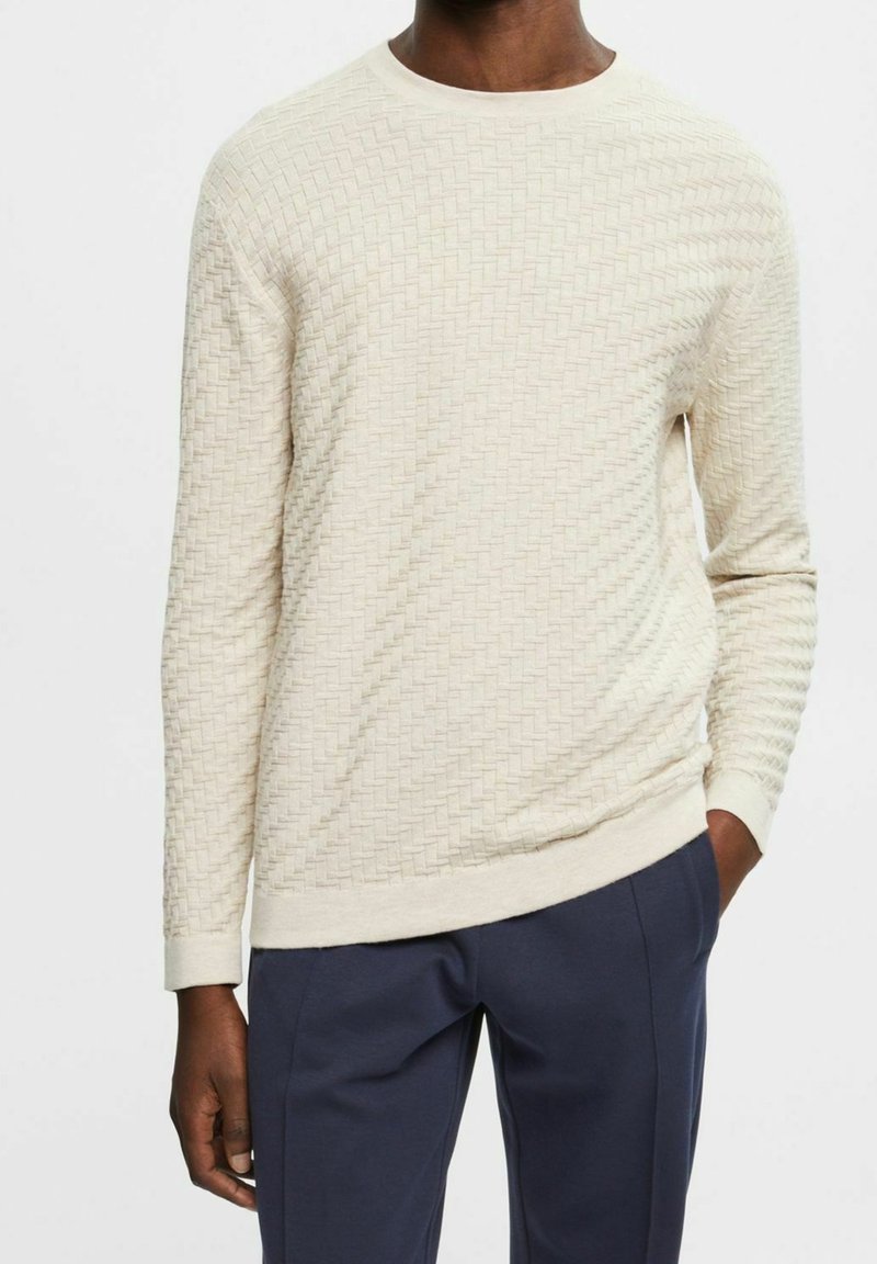 Homme portant un pull à manches longues texturé de couleur crème et un pantalon bleu marine, les mains relaxées le long du corps, sur un fond uni.