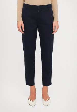 Nainen, jolla on yllään tummansiniset suoralahkeiset housut, beige pitkähihainen paita ja valkoiset kärjestä teräväkärkiset korkokengät, seisomassa yksivärisen taustan edessä.