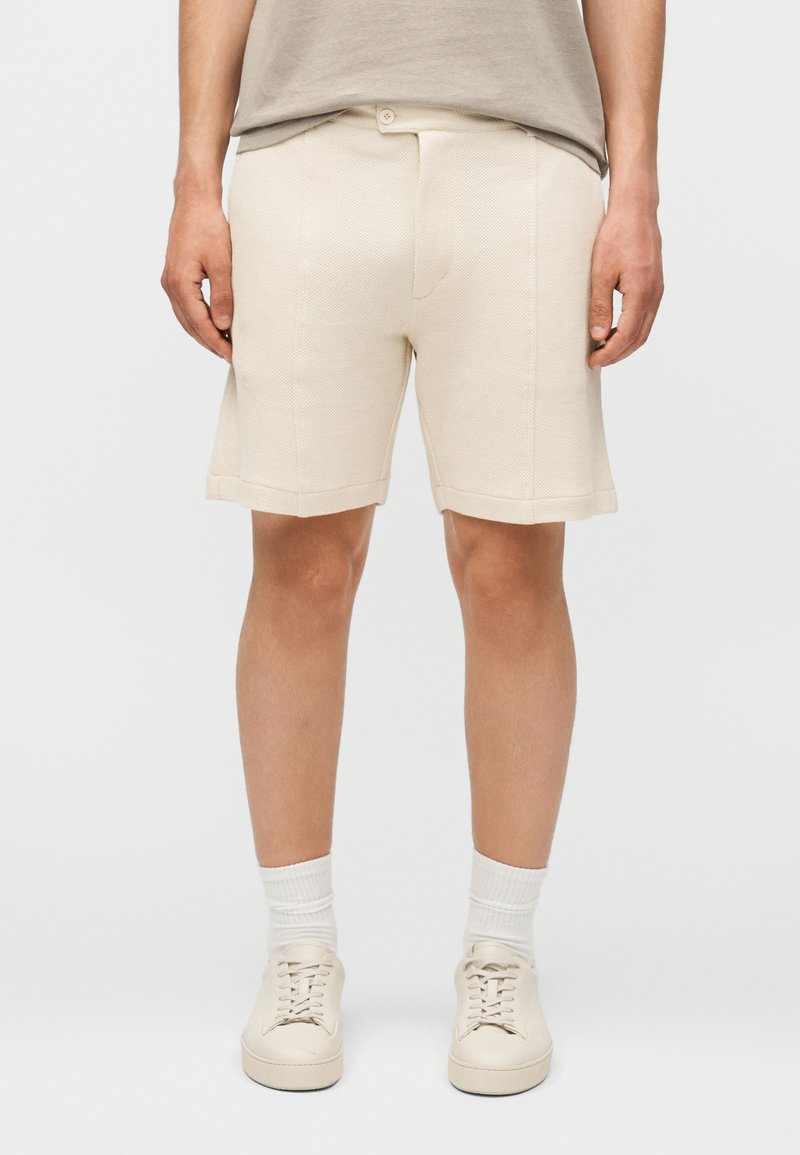 Άνδρας που φοράει μπεζ υφασμάτινα σορτς μέχρι το γόνατο, μπεζ αθλητικά παπούτσια, λευκές κάλτσες και ένα ανοιχτό ταουπέ t-shirt, όρθιος μπροστά σε απλό φόντο.