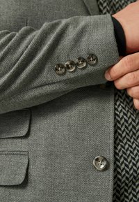 Grau strukturiertes Stoffblazer mit vier silbernen Knöpfen an den Ärmeln und einem kontrastierenden gemusterten Innenfutter. Figurbetonte Passform mit Seitentaschen.