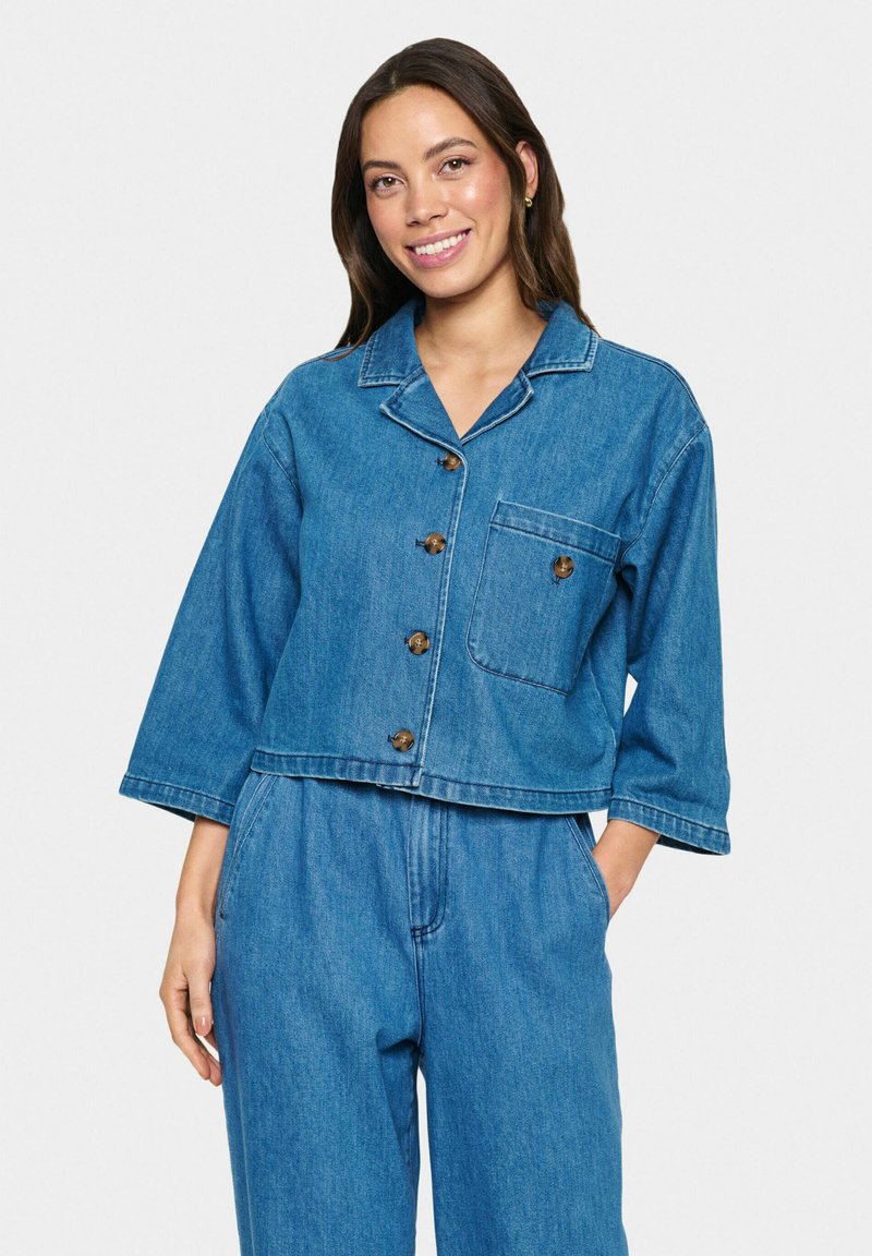 Denim jakna v svetlo modri barvi s skrajšanim dizajnom, gumbom spredaj, ovratnikom z izrezom in enim žepom na prsih. Ima dolge rokave in šivane detajle.