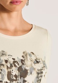 Cremefarbene Sweatshirt mit Rundhalsausschnitt, das ein strukturiertes Design in schwarzem und silbernem, abstraktem Muster auf der Vorderseite aufweist.