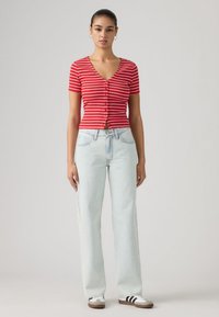 Κόκκινο και λευκό ριγέ πουκάμισο με κοντά μανίκια και κουμπιά, συνδυασμένο με ανοιχτό μπλε τζιν ψηλόμεσο και μπεζ αθλητικά παπούτσια με μαύρες ρίγες.