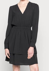 Svart klänning med djup V-ringning, långa ärmar och elastiskt midjeband. Flödande tyg med lagerdesign och knälång nederkant.