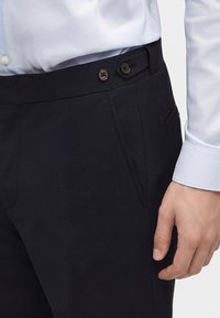 Άνδρας που φοράει μαύρο παντελόνι με κουμπιά ρύθμισης στο πλάι και λευκό πουκάμισο με κουμπωμένα μανίκια. Το χέρι είναι χαλαρό στο πλάι.