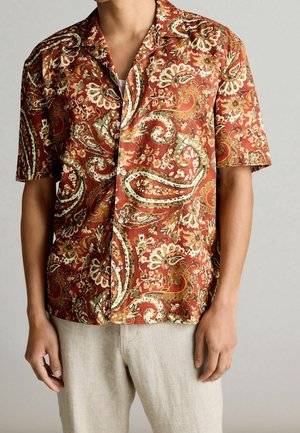 Mann trägt ein kurzärmeliges Hemd mit rotem und braunem Paisley-Muster und hellbeiger Hose vor einfachem Hintergrund.