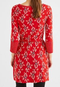 Punainen kukkakuvioinen mekko, jossa pitkät hihat, lehtien ja kukkien kuvio, selkeästi korostettu vyötärö ja pilkulliset yksityiskohdat hihoissa.