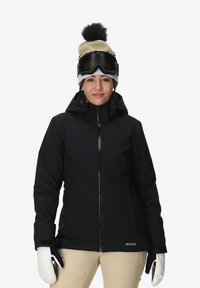 Giacca da sci nera con cappuccio, cerniera frontale, tasche laterali, indossata con guanti bianchi. Outfit completato con un berretto beige e occhiali da sci.