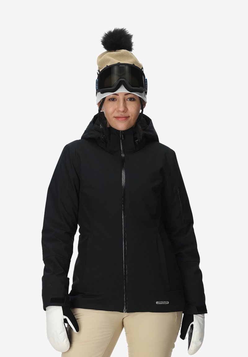 Giacca da sci nera con cappuccio, cerniera frontale, tasche laterali, indossata con guanti bianchi. Outfit completato con un berretto beige e occhiali da sci.