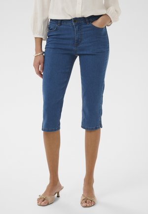 Frau trägt mittelhohe, hochgekürzte Jeans in mittlerem Blau mit mittlerer Taille und beigefarbene Sandalen mit Absatz und offenem Zehenbereich.