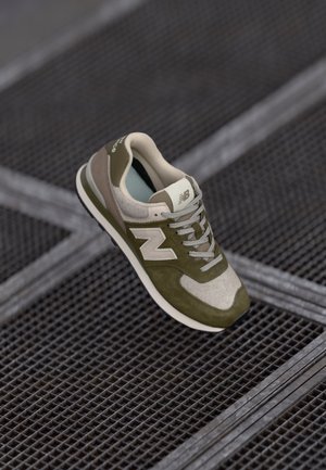 Olijfgroene en beige New Balance sneaker hangend boven een metalen rooster met kruislings patroon.