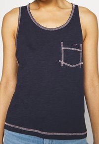Marineblauw tanktop met een zak. Kenmerkt zich door roze stiksels en een ronde halslijn. Gemaakt van een zachte, gestructureerde stof.