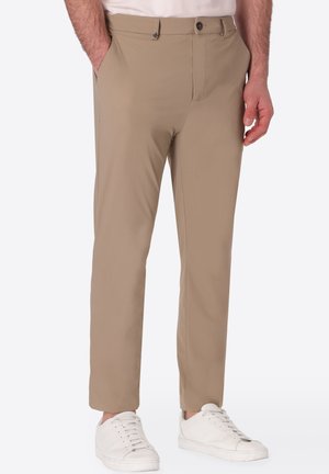 Uomo che indossa pantaloni beige a gamba dritta e scarpe da ginnastica bianche, in piedi con una mano in tasca davanti a uno sfondo semplice.