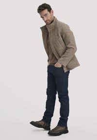 Braune Wolljacke mit hohem Kragen, fleecegefüttert und mit Seitentaschen. Getragen über einem braunen Pullover, kombiniert mit dunkelblauen Jeans und braunen Stiefeln.