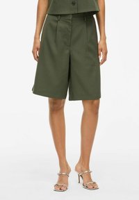 Pantaloni corti verde oliva su misura con una vestibilità rilassata, pieghe sul davanti e una superficie liscia. Indossati con sandali con tacco alto argentati.