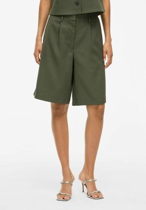 Olive green tailored shorts with a relaxed fit, pleats at the front, and a smooth texture. Worn with silver high-heeled sandals.