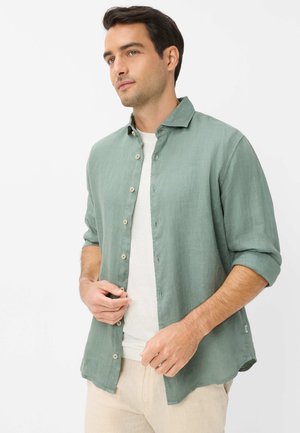 Mann mit dunklem Haar, der ein olivgrünes Hemd mit hochgekrempelten Ärmeln über einem weißen T-Shirt und beiger Hose trägt, vor weißem Hintergrund stehend.