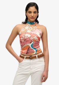 Zijden haltertop met een kleurrijk bloemen- en pauwenpatroon, voorzien van een blauwe halsafwerking. Gecombineerd met witte broek en een gevlochten bruine riem.