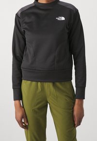 Musta huppari, jossa on korkea kaulus, teksturoitu kangas, roikkuvat olkapäät ja logo vasemmassa rinnassa. Yhdistetty oliivinvihreisiin housuihin.