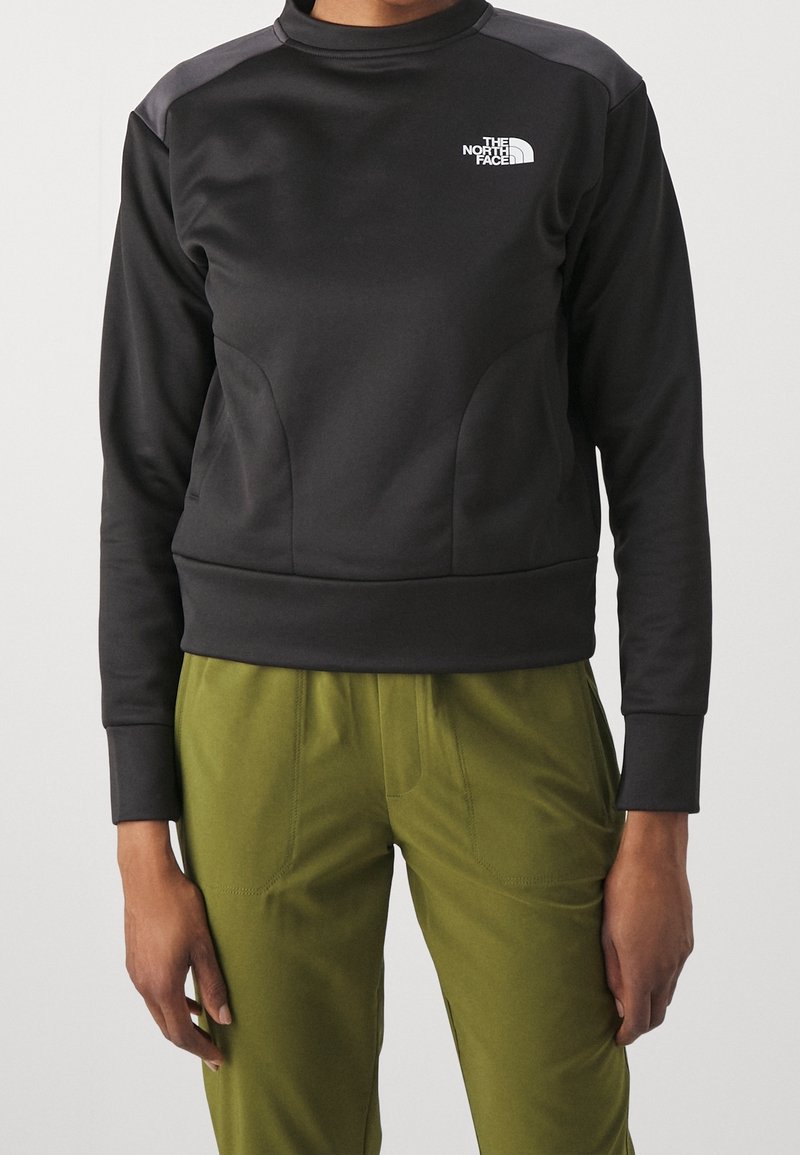 Musta huppari, jossa on korkea kaulus, teksturoitu kangas, roikkuvat olkapäät ja logo vasemmassa rinnassa. Yhdistetty oliivinvihreisiin housuihin.