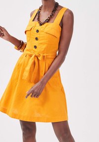 Robe en lin jaune avec une jupe évasée, décolleté carré, deux poches poitrail, six boutons noirs et une ceinture nouée à la taille.