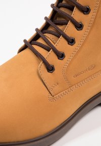 Botte en daim beige avec lacets marron, œillets hexagonaux et logo géométrique. Présente une texture lisse et un bout arrondi.