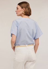 Blusa a rayas azul claro con mangas cortas, con un cuello redondo adornado con cuentas blancas decorativas. Usada con pantalones blancos de cintura alta.