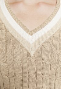 Beige stickad tröja med V-hals, med krämfärgade ränder och flätad textur. Närbild som betonar materialdetaljer och design.