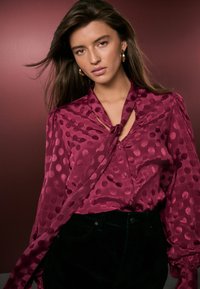 Blusa di seta color bordeaux con collo a fiocco, decorata con grandi pois rosa, con bottoni sul davanti e maniche arricciate. Indossata con pantaloni scuri.
