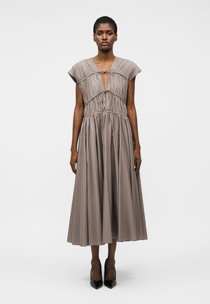 Kobieta w bezrękawnikowej sukience midi w kolorze taupe z marszczeniami i wiązaniami na biuście, w czarnych szpilkach z ostrym noskiem, stojąca twarzą do przodu.