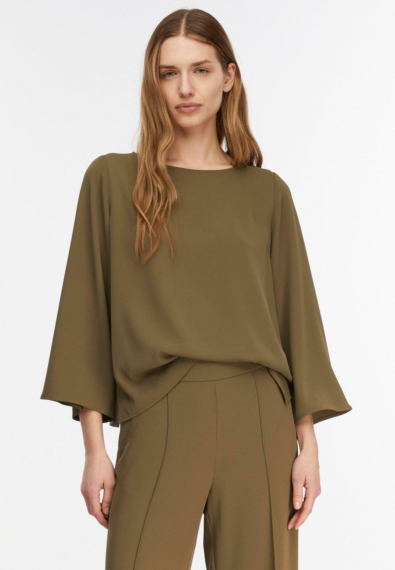 Femme aux longs cheveux portant un chemisier ample vert olive à manches larges et un pantalon taille haute assorti, debout devant un fond blanc.