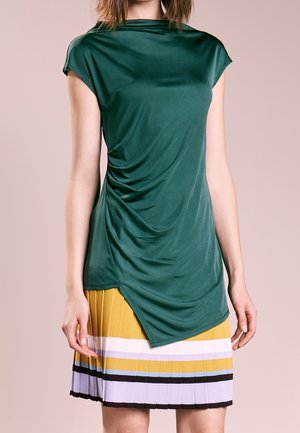 Femme portant un haut à manches courtes vert foncé avec drapé sur le côté et ourlet asymétrique sur une jupe plissée avec des rayures jaunes, blanches, noires et violet clair.