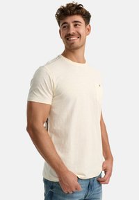 Cremefarbenes T-Shirt mit kurzen Ärmeln aus leichtem Stoff, ausgestattet mit einer linken Brusttasche und Kontrastnähten an den Schultern.